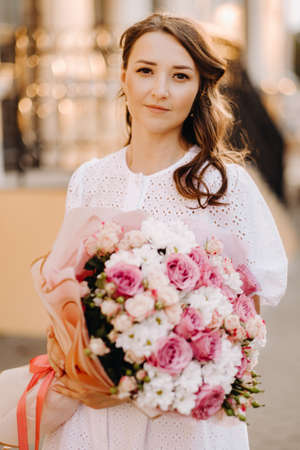 A happy woman in a white dress at sunset with a bouquet of flowers in the city.の写真素材