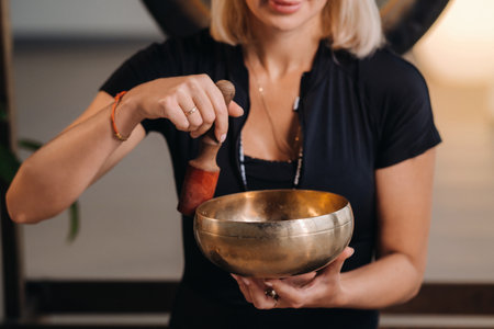 A woman plays a Tibetan singing bowl while sitting on a yoga mat against the background of a gongの写真素材