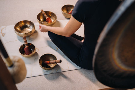 A woman sits in a lotus position next to Tibetan bowls, sitting on a yoga mat against the background of a gong.の写真素材