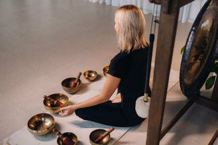 A woman sits in a lotus position next to Tibetan bowls, sitting on a yoga mat against the background of a gongの写真素材