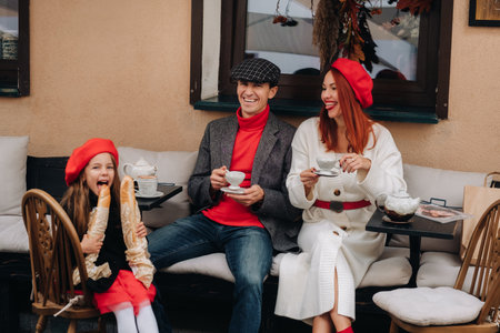 A stylish family of three is sitting at a table outside in a cafe and drinking coffee. Dad, mom and daughter in the autumn cityの写真素材