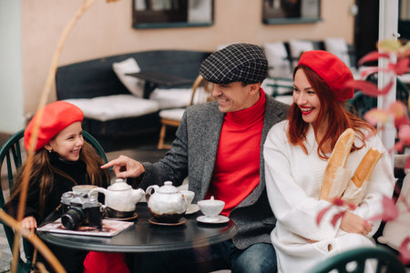 A stylish family gathered together in a cafe on the street. Mom, dad, little daughter drink tea, eat cakes. They are happy together. The concept of a happy family dinnerの写真素材