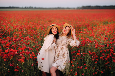 Two girlfriends in dresses and a hat in a poppy field in summer at sunsetの写真素材