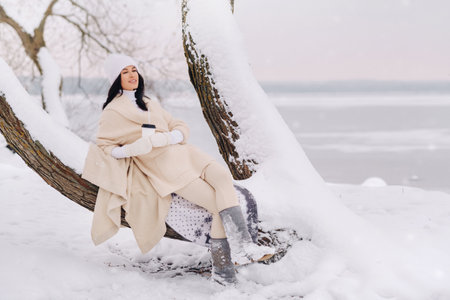 A beautiful girl with a beige cardigan and a white hat enjoying drinking tea in a snowy winter forest near a lakeの写真素材