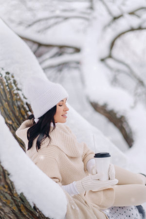 A beautiful girl with a beige cardigan and a white hat enjoying drinking tea in a snowy winter forest near a lakeの写真素材