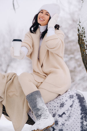 A beautiful girl with a beige cardigan and a white hat enjoying drinking tea in a snowy winter...