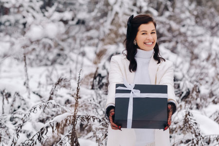 A stylish woman with a white suit with a New Years gift in her hands in a winter forest. A girl in nature in a snowy forest with a gift boxの写真素材