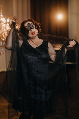 An elegant adult woman in a black cocktail dress and mask poses in the interior. Fashionable female...
