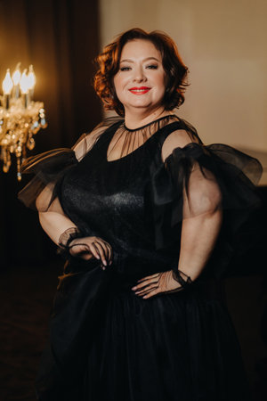 An elegant adult woman in a black cocktail dress poses in the interior. Fashionable portrait of a...