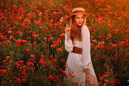 a girl in a white dress and hat stands in a field with poppies at sunset and holds a poppy flower in her hand.の写真素材