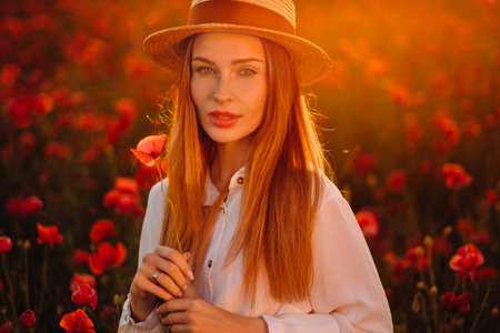 a girl in a white dress and hat stands in a field with poppies at sunset and holds a poppy flower in her handの写真素材