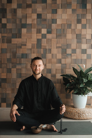 Portrait of a young man in a black kimano sitting in a lotus position on a gym mat in the interior.