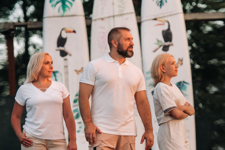 A family of three stands against a backdrop of surfboards at sunset.の写真素材