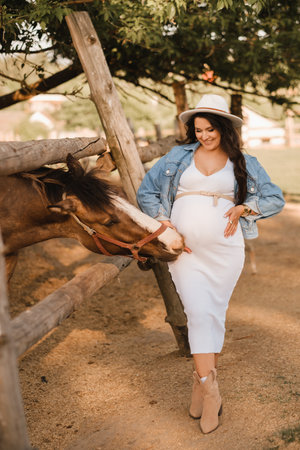 Stylish pregnant woman in a white hat in the countryside near a horseの写真素材