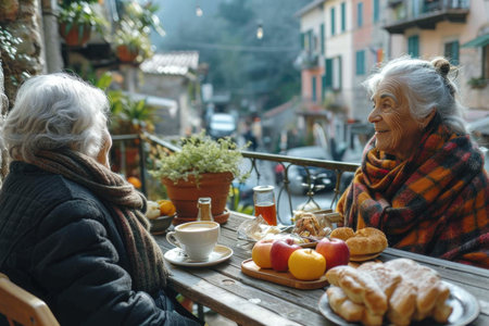 Two elderly female friends are sitting at an outdoor table with cups of coffee and smiling at the camera.の素材