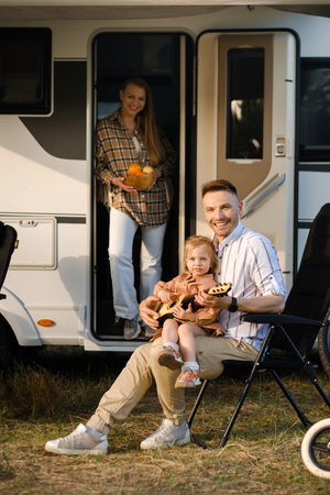 A family takes pleasure in a day spent outdoors by their camper van, with the father holding their child and the mother bringing refreshments, all surrounded by greenery and sunshine.の写真素材