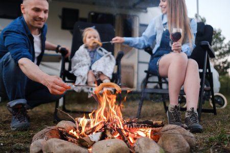 A family gathers around a campfire in the outdoors, grilling sausages on sticks as they enjoy food and drink together. The atmosphere is warm and relaxed, perfect for bonding.の写真素材