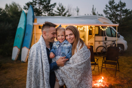 A family of three embraces warmly by a campfire, wrapped in blankets. Their camper van and surfboards set the backdrop for a joyful evening outdoors surrounded by nature.の写真素材