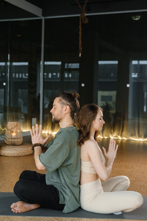 Two individuals sit back to back on yoga mats, practicing mindfulness and meditation in a tranquil indoor studio as soft lighting creates a calming atmosphere.の写真素材
