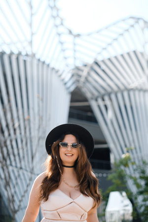 A woman with long hair and sunglasses stands in front of unique architectural design. She wears a fashionable outfit and a wide-brimmed hat, enjoying the sunny day outdoors.の写真素材