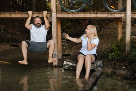 Two friends are having fun by the water on a sunny day. One is playfully hanging from a wooden...