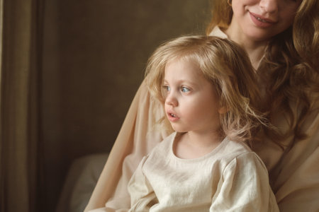 A mother holds her young daughter as they sit together in a warmly lit room. The child gazes...