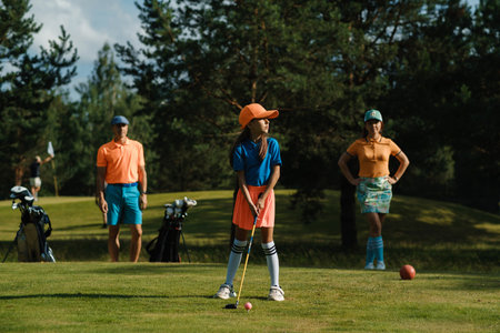 A young girl prepares to hit a ball on a sunny golf course surrounded by her parents, playing a family game of golf on a weekend morningの写真素材