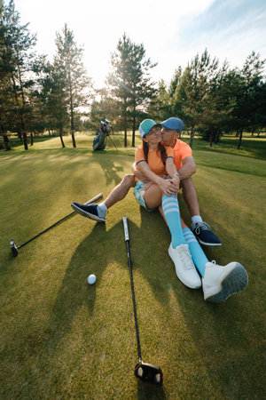 A couple in colorful clothes sits on a golf course and kisses on a sunny day, surrounded by trees and golf equipmentの写真素材