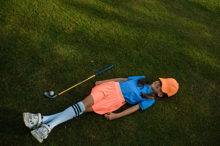 A young girl in a bright sports outfit is relaxing on the green grass on a sunny day at the golf course, tired after half a workoutの写真素材