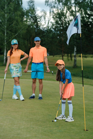 Family of three enjoying a fun day of golfing, with a young girl preparing to putt as her parents...
