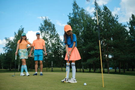 Young girl prepares to putt on a golf course while parents watch nearby on a sunny day with bright...