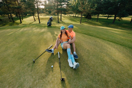 A couple relaxes on the golf green, wearing colorful outfits. They sit back-to-back, enjoying each other's company after a playful round of golf in the afternoon sun.の写真素材