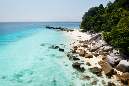 Beautiful beach scene featuring turquoise waters, rocky outcrops, and sandy shores. People enjoy swimming and sunbathing under the bright sun surrounded by lush greenery.の写真素材