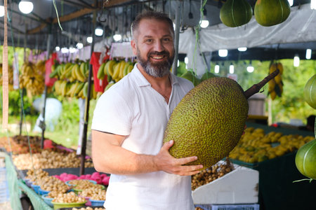 A man with a beard smiles while holding a large jackfruit in a lively outdoor market filled with...