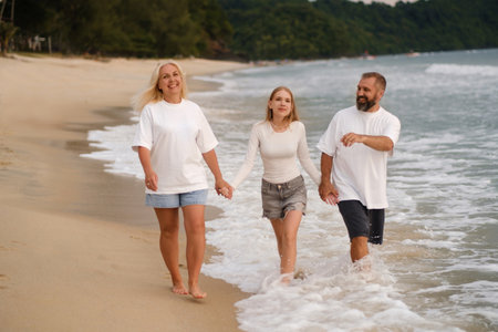 Two adults and a teenager walk hand in hand along a sandy beach while waves crash gently at their feet. The sun sets in the background, creating a warm atmosphere.の写真素材