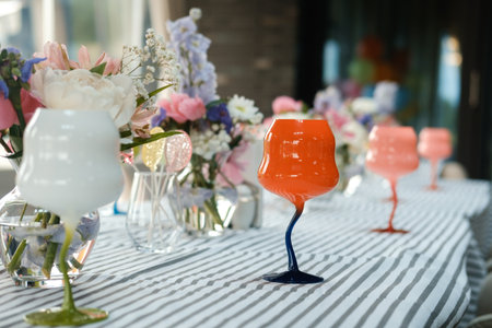 Vibrant colored glasses are arranged on a striped tablecloth alongside fresh flowers in vases, creating a lively atmosphere for a gathering in a well-lit indoor area.の写真素材