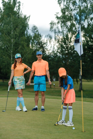 On a sunny day at a golf course, a young girl in a blue shirt and orange cap prepares to make a putt, while her family watches her closely, all dressed in vibrant golf attire.の写真素材