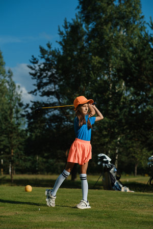 A young girl in a bright blue shirt and orange skirt focuses on her swing while practicing golf on a sunny day. Lush trees and equipment create a vibrant atmosphere on the course.の写真素材