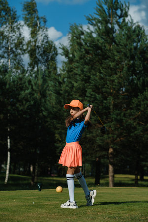 A girl swings a golf club with focus, wearing a bright outfit, while practicing on a lush course under a clear blue sky, showcasing her dedication to the sport.の写真素材