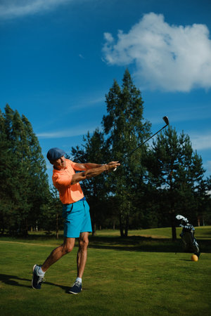 A male golfer dressed in bright attire takes a powerful swing on a lush green golf course during a sunny day. Trees surround the area, enhancing the tranquil atmosphere.の写真素材