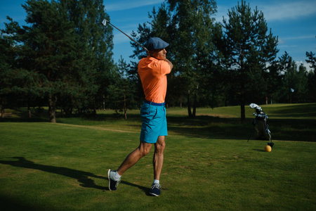 A man in an orange shirt and blue shorts is executing a golf swing on a well-maintained course. Trees stand tall in the background under a clear sky.の写真素材