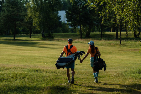 A man and woman walk side by side on a golf course, sharing smiles and laughter while carrying their golf clubs and bags, enjoying a sunny afternoon surrounded by trees.の写真素材