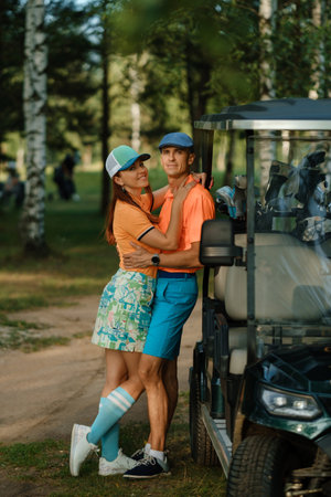A couple shares a warm embrace by a golf cart in a green park, both wearing colorful golf outfits. Sunlight filters through the trees, creating a cheerful atmosphere.の写真素材