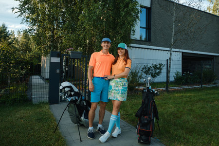 A couple smiles broadly while dressed in vibrant golf outfits, standing outside a modern house. They are ready to enjoy a day of golfing on a sunny afternoon, surrounded by nature.の写真素材