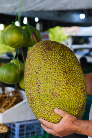 A person holds a large jackfruit at a bustling market filled with colorful fruits. The scene...
