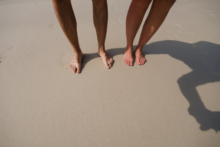 Two people are standing barefoot on a light sandy beach, enjoying the warm sunlight and soft waves playing at their feet during a relaxing summer day by the sea.の写真素材