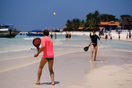 Two individuals play beach tennis on a sunny day, surrounded by a lively beach scene with people walking and boats in the distance under a clear blue sky.の写真素材