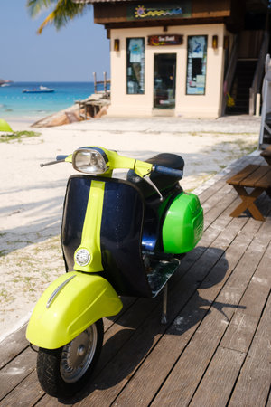 A bright green and yellow scooter stands on a wooden deck overlooking a sandy beach and turquoise water. Palm trees sway nearby, enhancing the tropical atmosphere under the sun.の写真素材