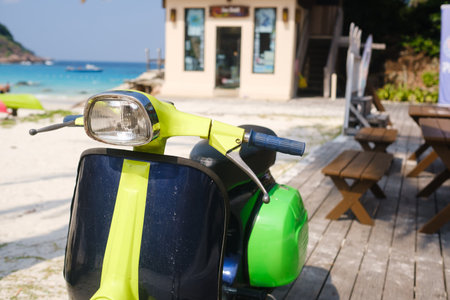 A colorful scooter stands parked on a wooden deck, overlooking the sandy beach and turquoise water. Beach activities can be seen in the background, creating a relaxing atmosphere.の写真素材