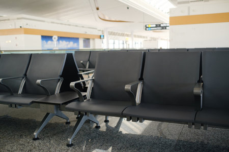 Empty black chairs are arranged in a spacious airport terminal. Natural light brightens the area, and clear boarding signs are visible in the background, indicating a calm travel atmosphere.の写真素材